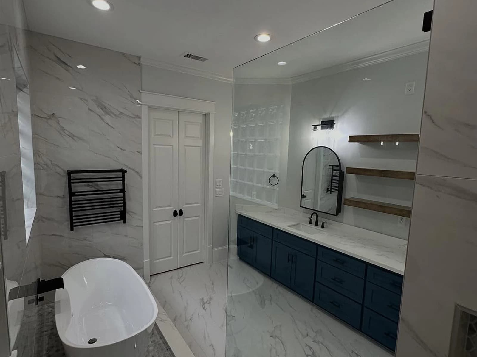 Finished bathroom remodel with freestanding tub, glass shower, blue vanity, floating shelves, and marble-look tile