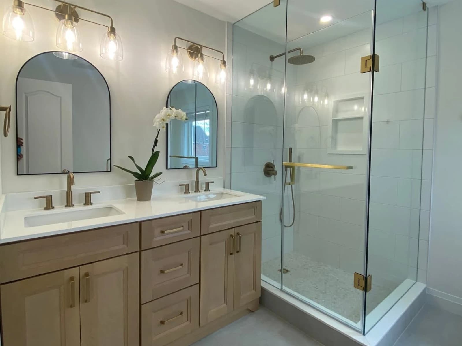 Bathroom remodel with wood vanity, twin mirrors, glass shower, brass hardware, and pale floor tile
