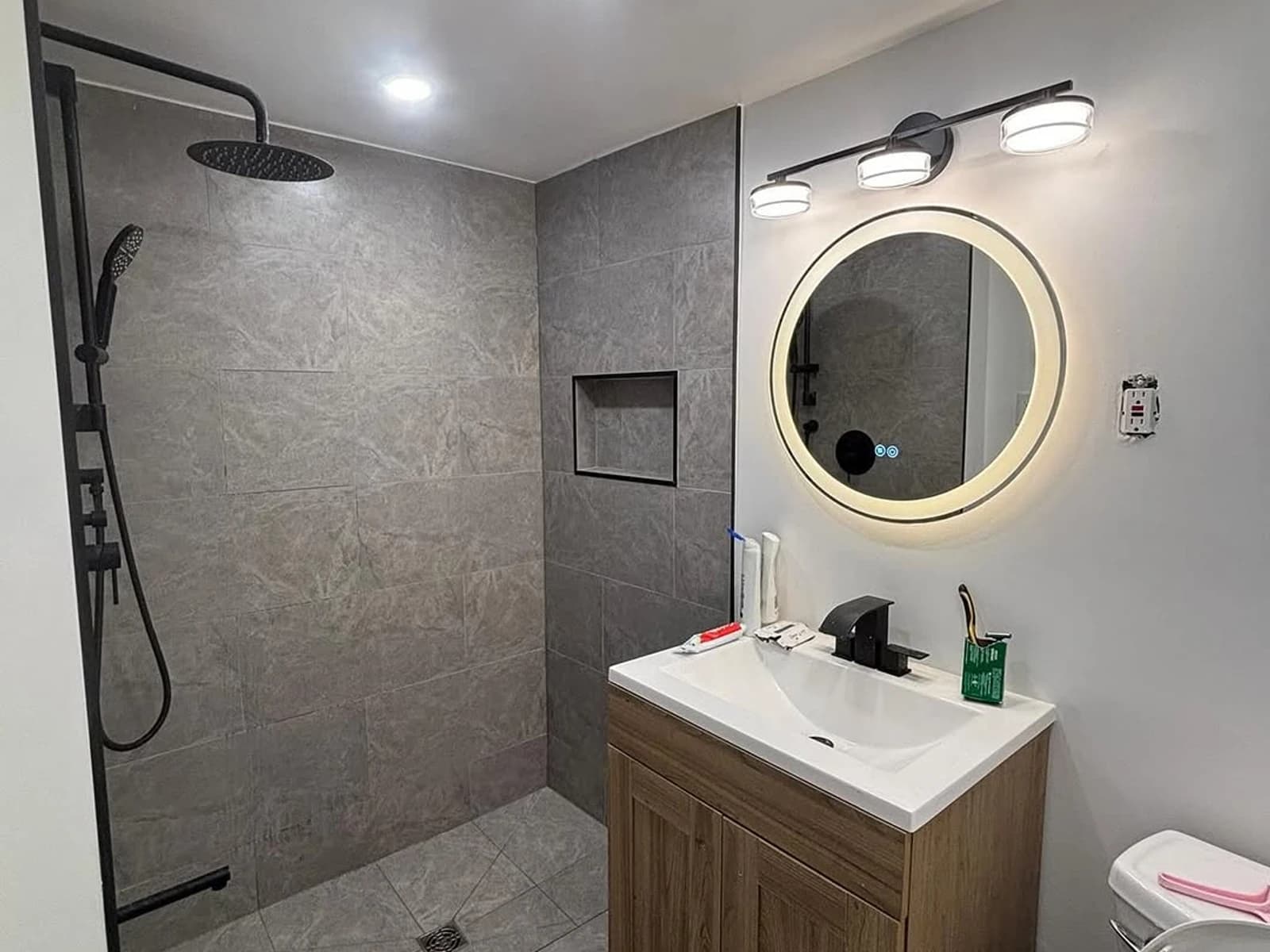 Gray tile walk-in shower with wood vanity, round mirror, and black fixtures